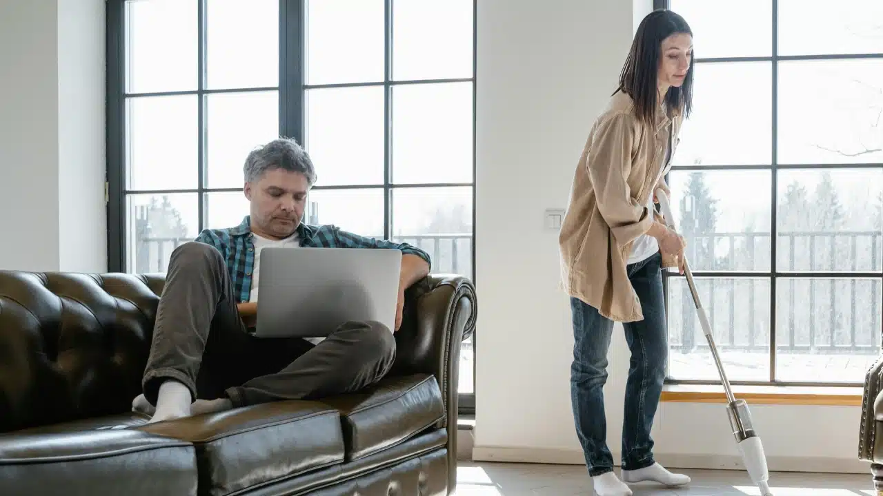 A man uses a laptop on a leather sofa while a woman vacuums the floor nearby.