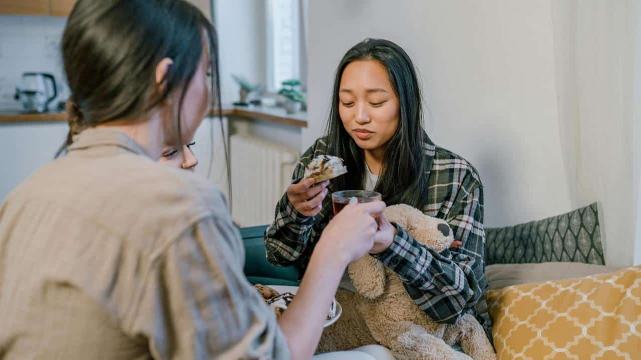 A woman eating a snack and drink while sitting on a couch with another person and a child.