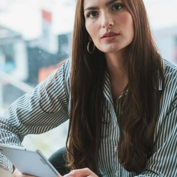 A woman with long brown hair wearing a striped shirt, holding a tablet and looking attentively