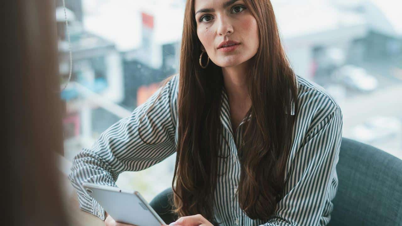 A woman with long brown hair wearing a striped shirt, holding a tablet and looking attentively