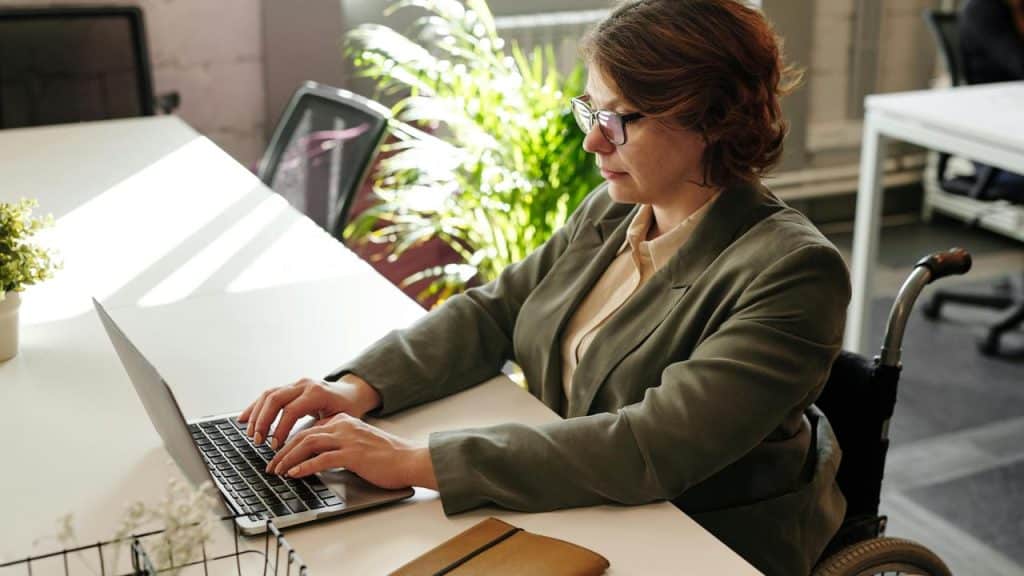 A woman in a wheelchair typing on a laptop at a desk in a sunlit office.