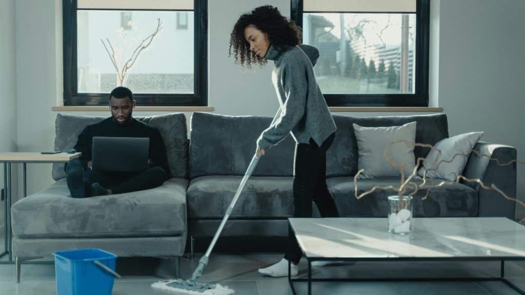 A woman mops the floor while a man sits on the sofa using a laptop.