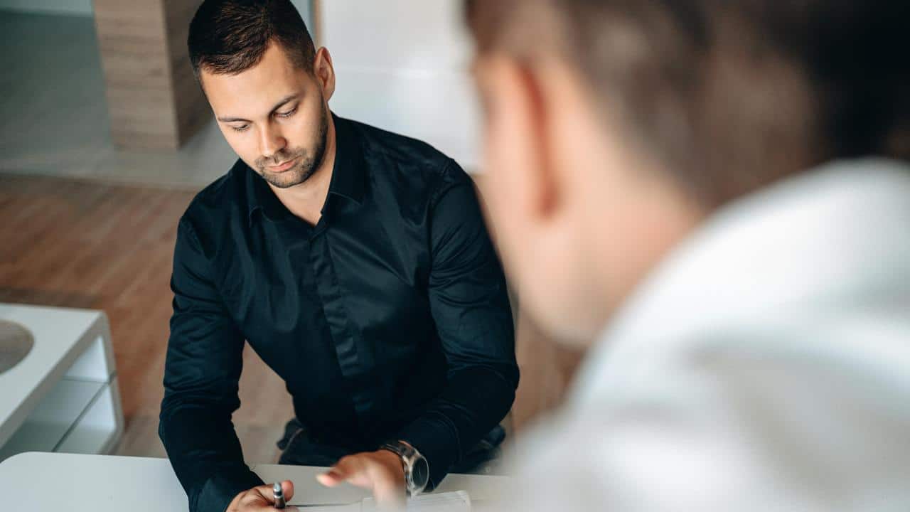 A person in a black shirt signing a document at a table with another person nearby.