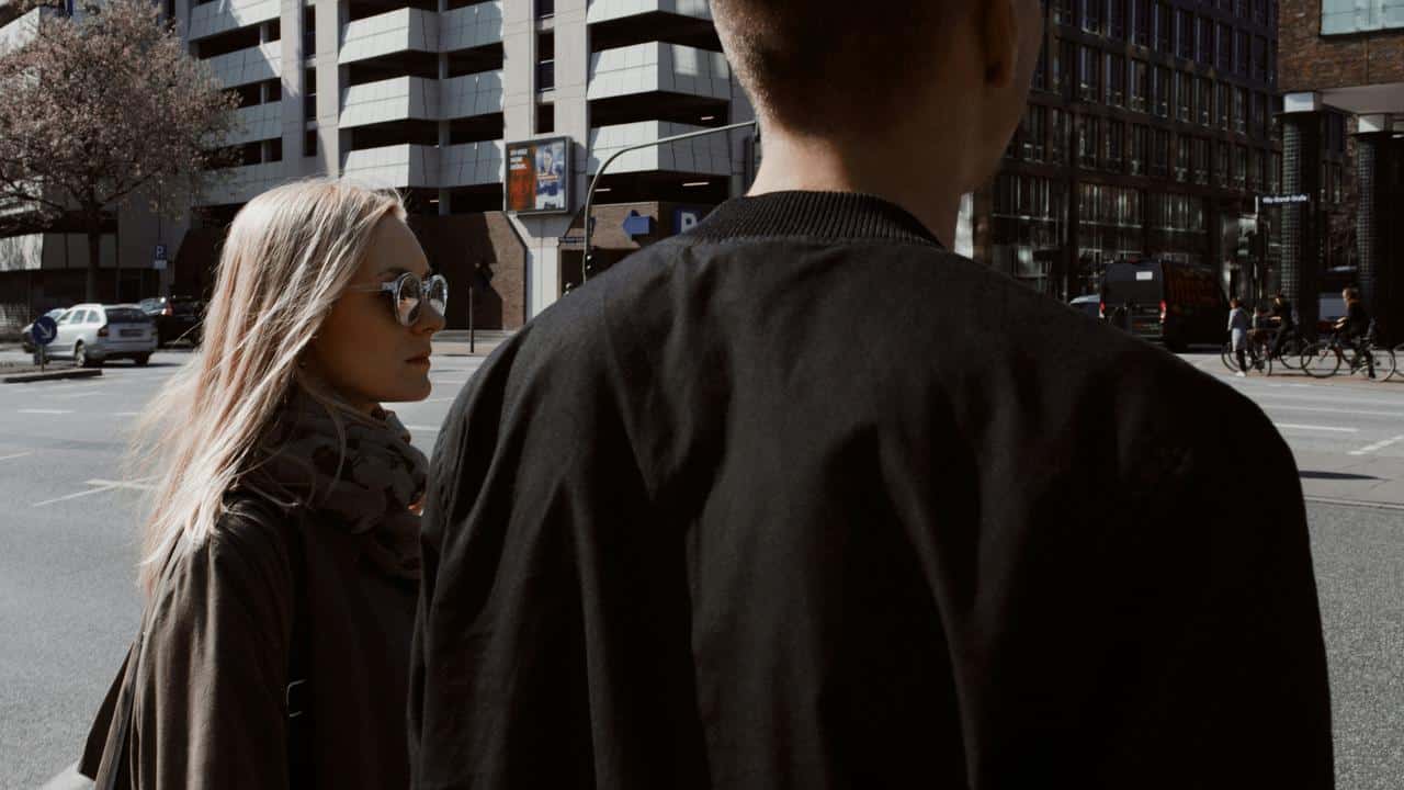 A couple standing at a city street crossing, viewed from behind.