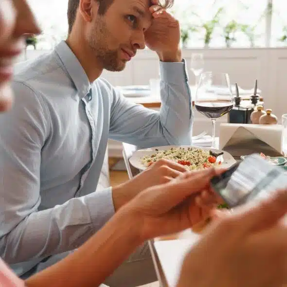 A couple at a restaurant table with food and wine, one person checking a phone.