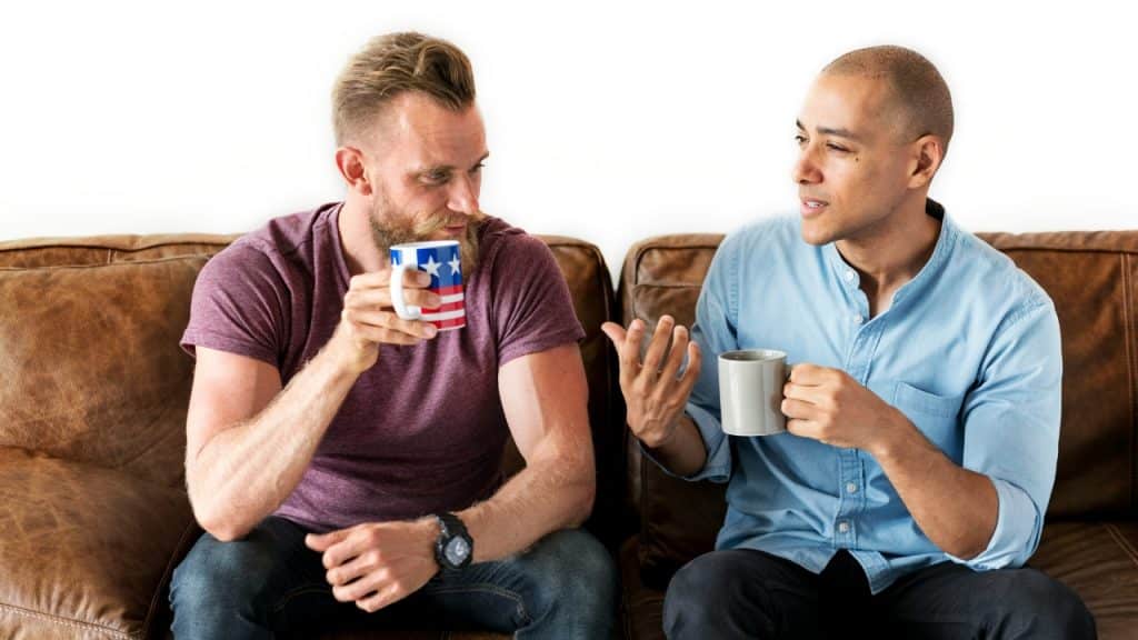 Two men sit on a leather sofa talking while holding coffee mugs.