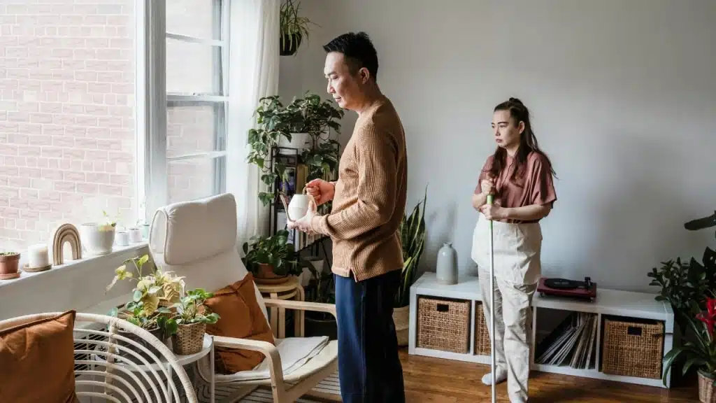 A man waters a plant by a window while a woman stands nearby holding a mop.