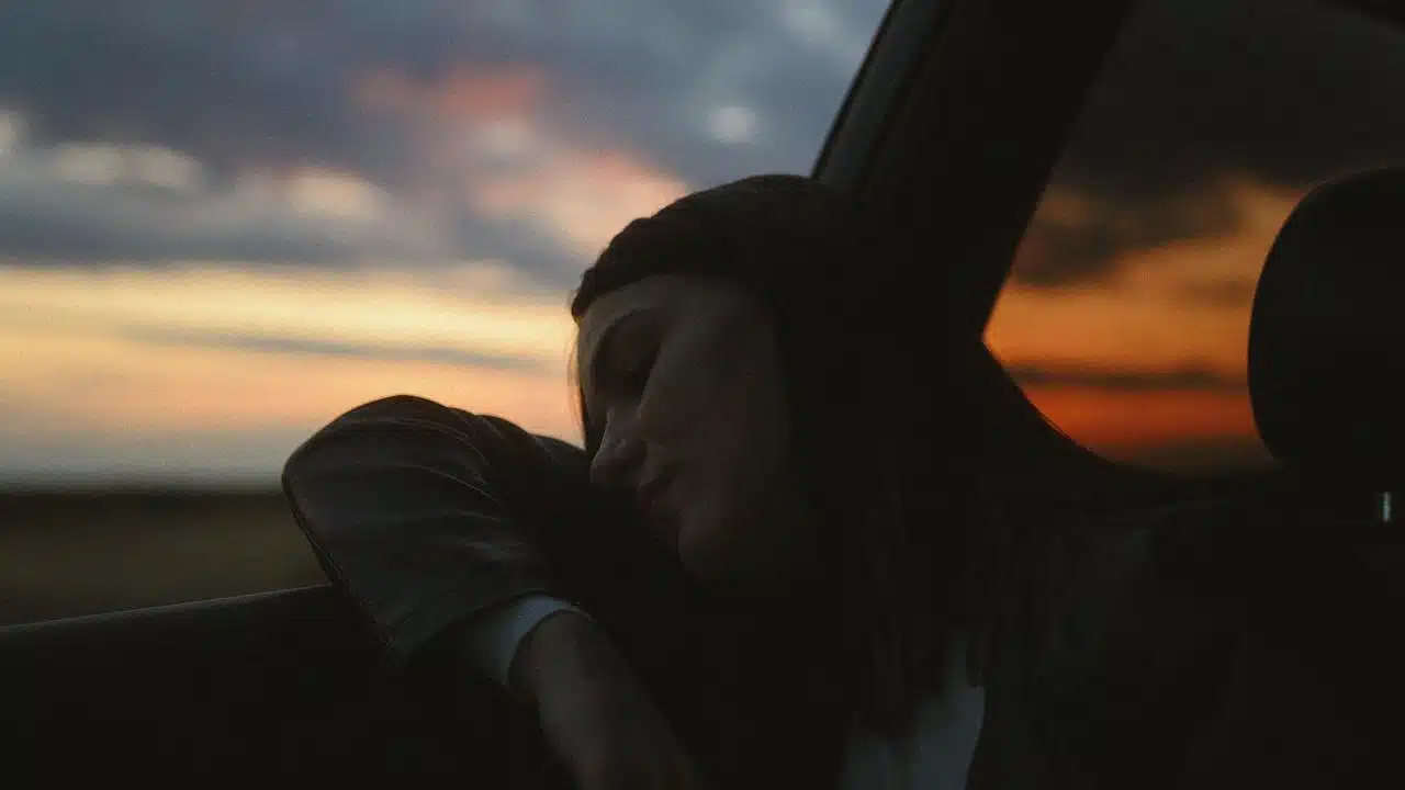 A woman resting her head against a car window at sunset.