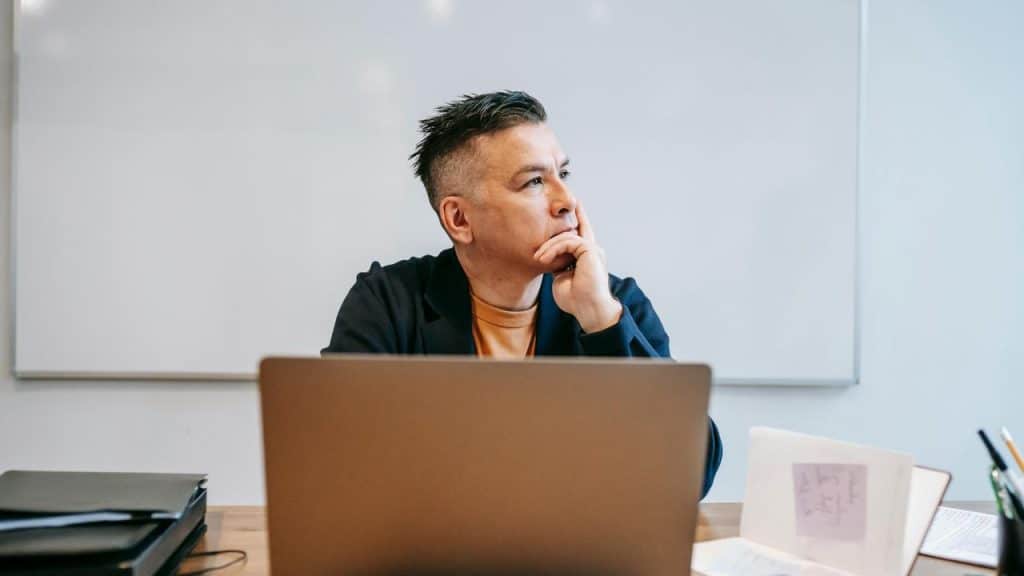 A man sitting at a desk with a laptop, looking away thoughtfully.