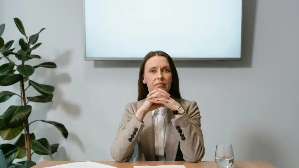 A serious woman in a blazer sitting at a desk with hands clasped.