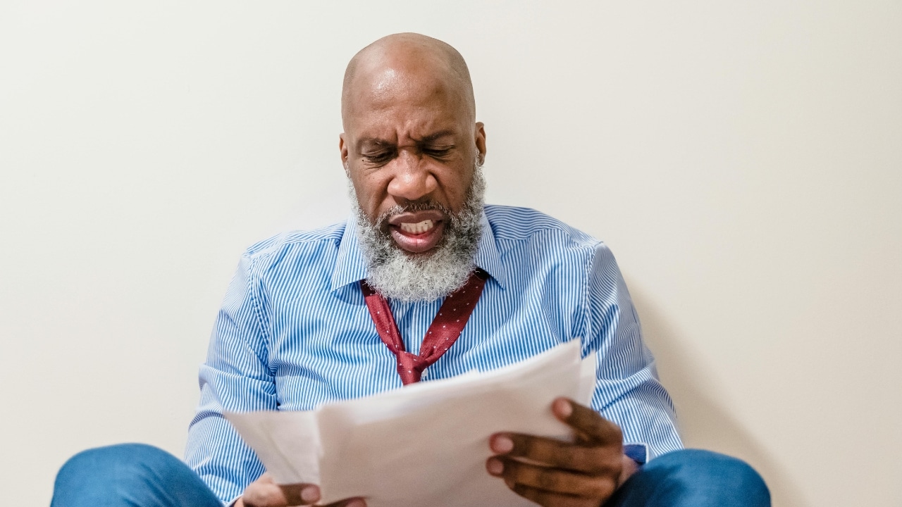 A man with a grey beard sits on the floor looking at papers with frustration.