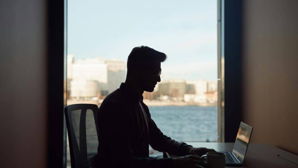 A man working on a laptop by a window, silhouetted against a cityscape and water view.