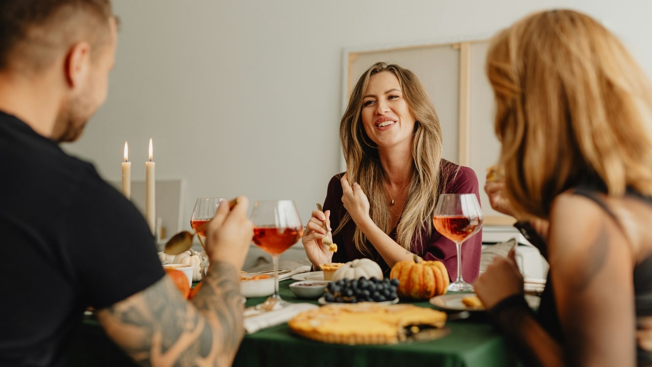 People sit around a dinner table with wine and pumpkins, smiling and talking together.