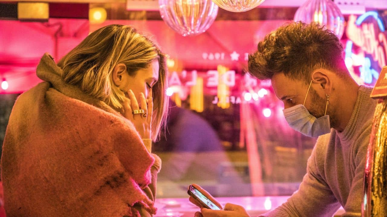 A man in a face mask looks at a smartphone while a woman sits nearby.