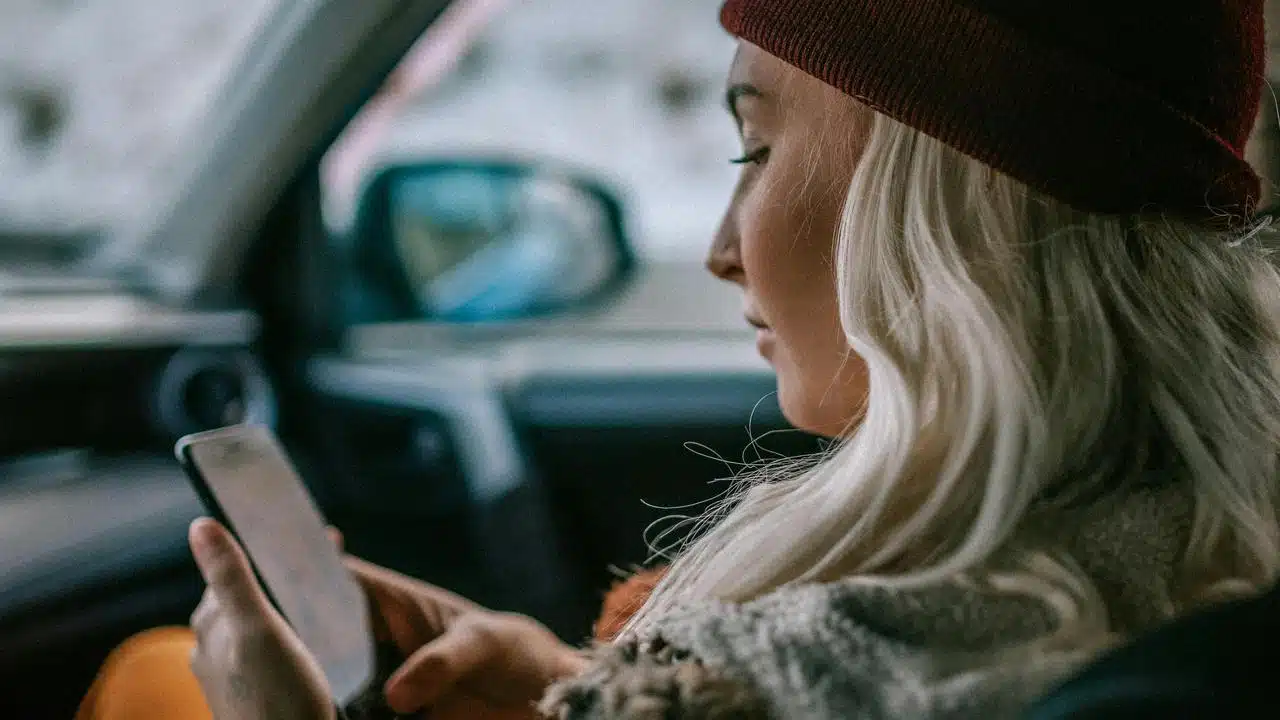 A woman wearing a red beanie looks at her phone while sitting in a car.