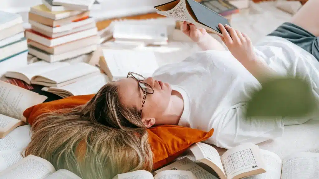 A woman lying on the floor surrounded by books while reading.