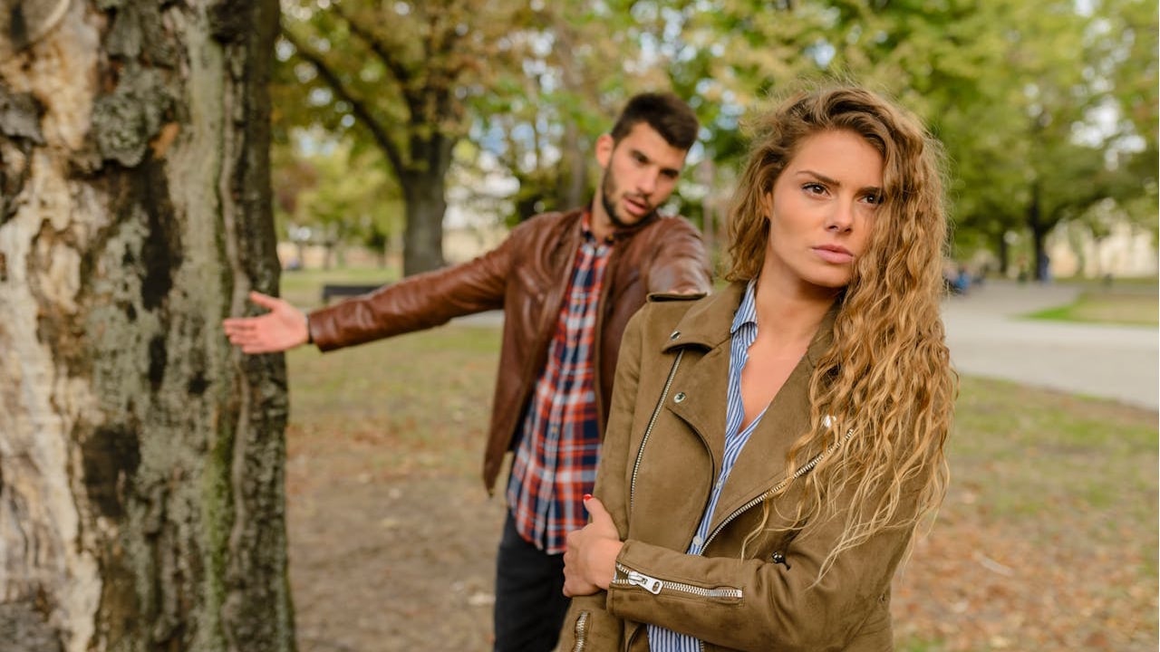 A woman with long wavy hair, wearing a brown jacket, is looking directly at the camera while a man gestures in the background near a tree.