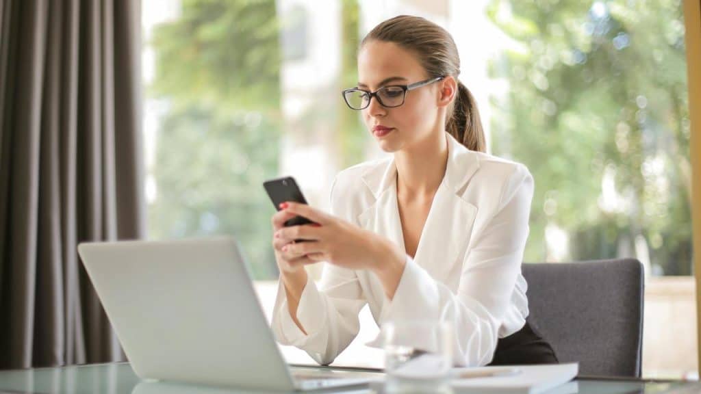 A woman wearing glasses sitting at a desk using her phone beside a laptop.