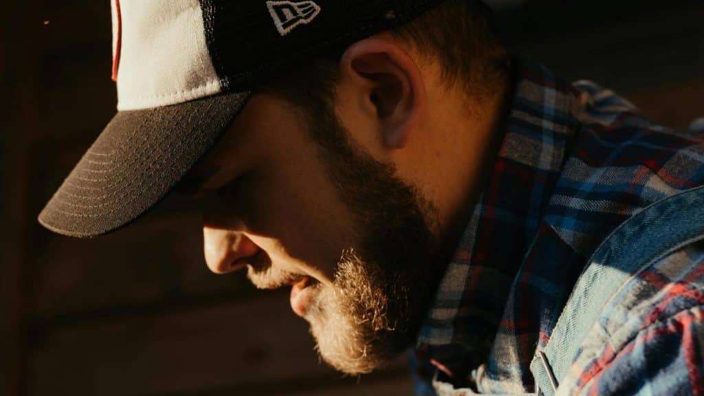 A bearded man wearing a cap looks downward in warm light.