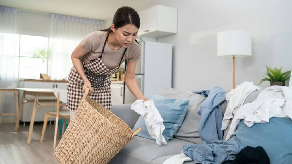 A woman in an apron gathers clothes from a sofa into a large wicker laundry basket.