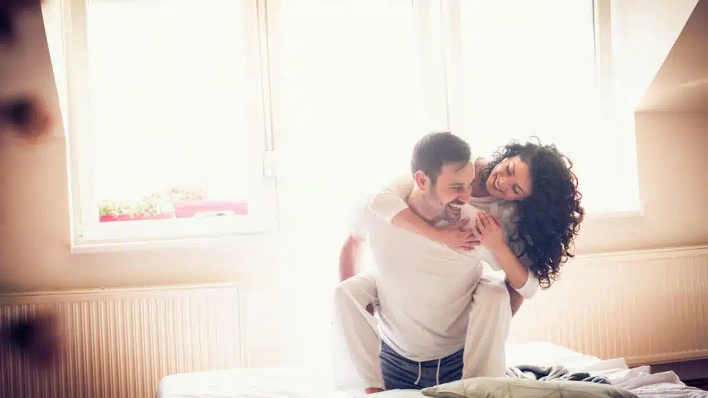 A man in white pajamas is giving a piggyback ride to a woman with curly hair on a bright bed in front of a large window.