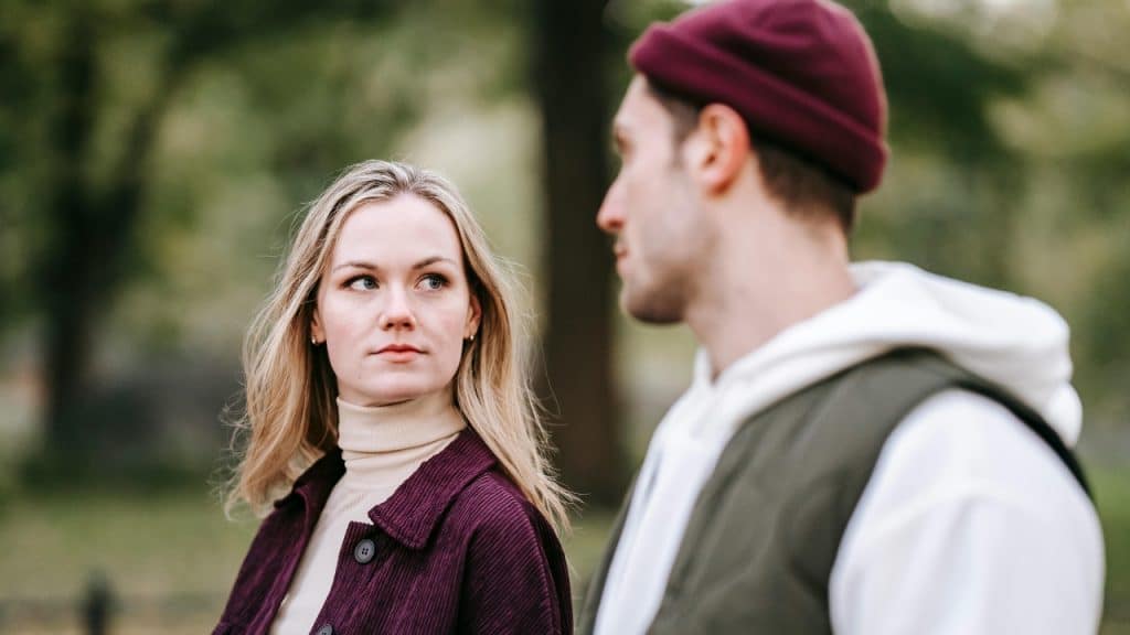 A blonde woman in a purple jacket looks toward a man wearing a maroon beanie.