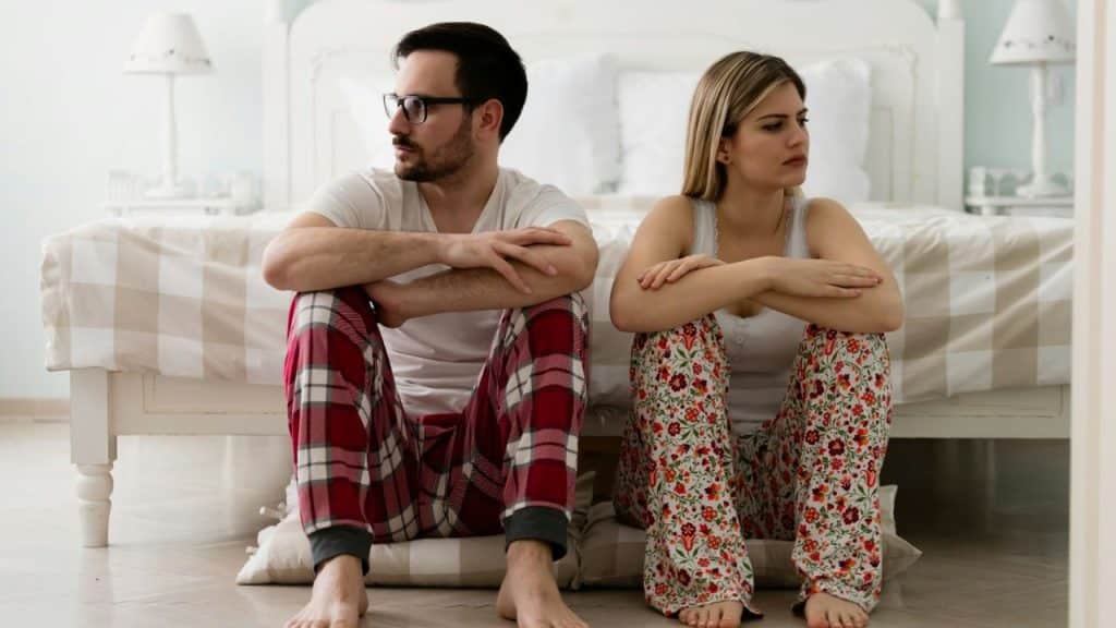 A man and woman sit on the floor by a bed, looking in opposite directions.