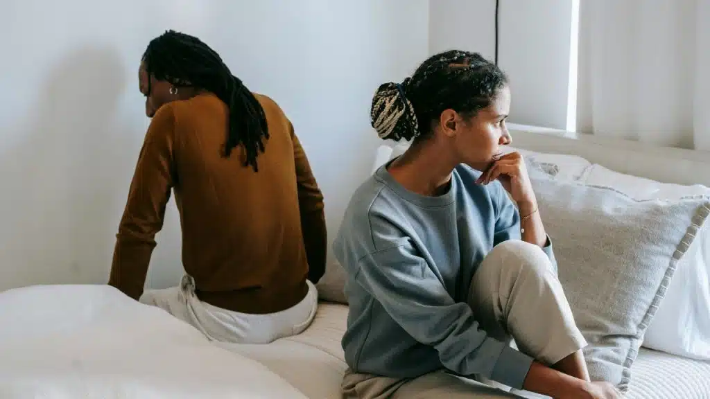Two people sit on a bed facing away from each other with somber expressions.