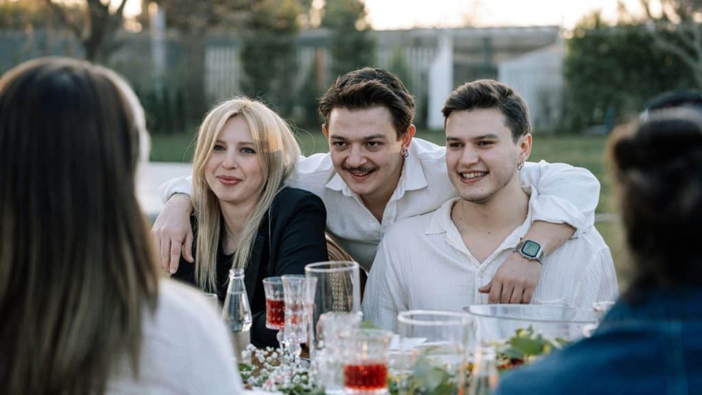 Three people smile while sitting at an outdoor table set with glasses and greenery.