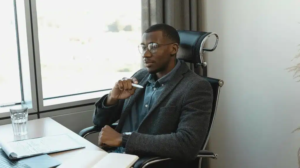 A man with glasses sitting thoughtfully at a desk by a window.