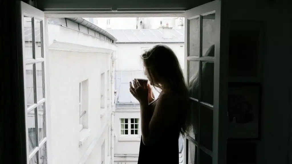 A woman standing by an open window holding a cup.