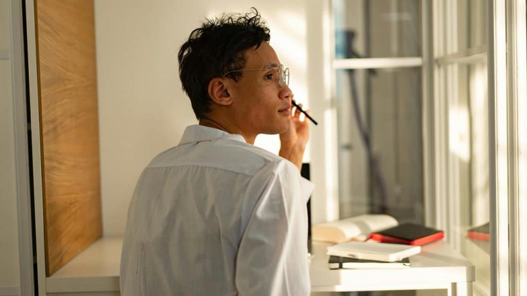 A man holding a pen near his face while looking out a window, appearing thoughtful.