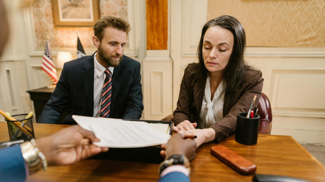 A man and woman in business attire sit at a desk while a document is presented.