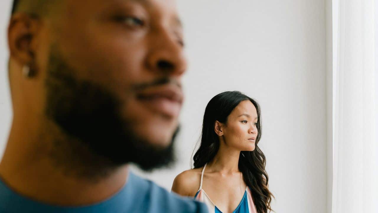 A woman with long dark hair looks toward a window behind a blurred man’s face.