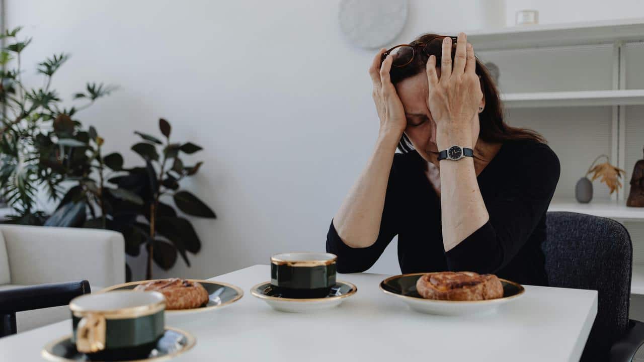 A person sitting at a table with pastries and coffee, holding their head in stress.