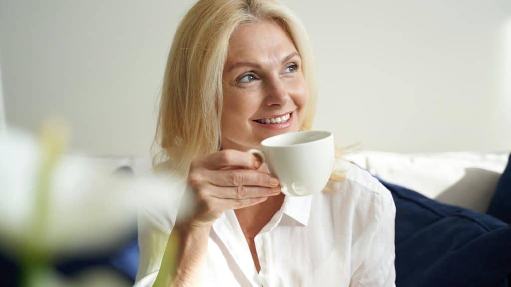 A woman in a white shirt smiling while holding a cup of coffee.