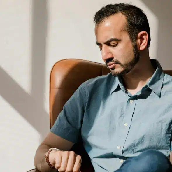 A man sitting in a leather chair, looking down thoughtfully in warm sunlight.