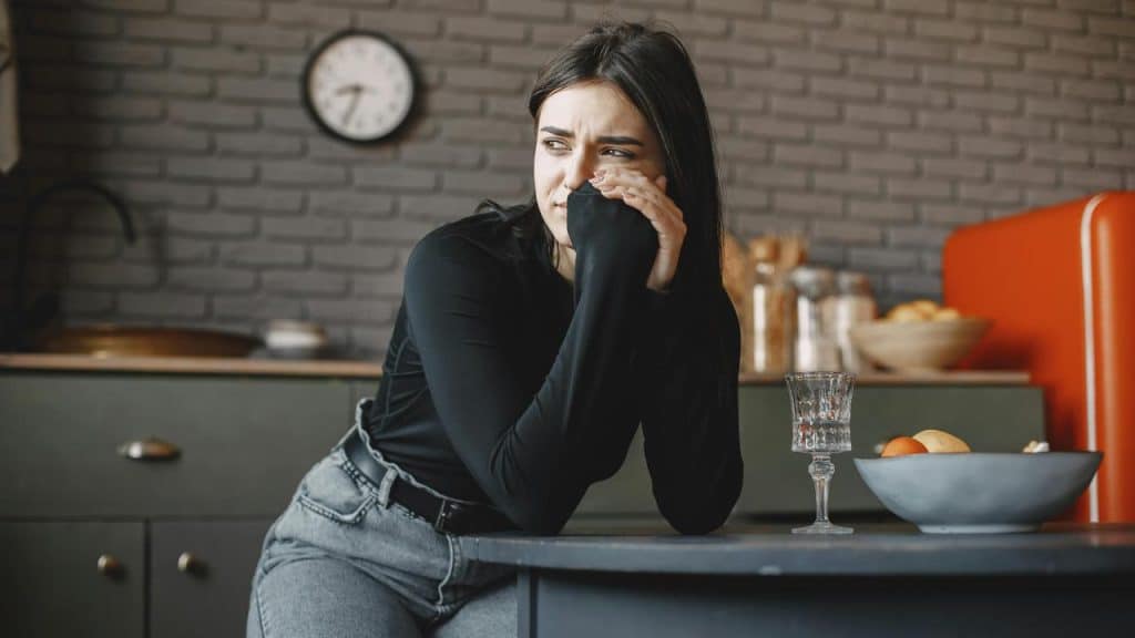 A worried woman wiping her eye while leaning on a kitchen counter.