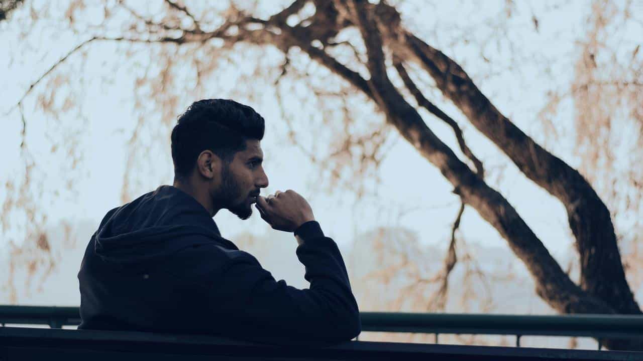 A man in a dark hoodie sitting on a bench outdoors, looking thoughtful.