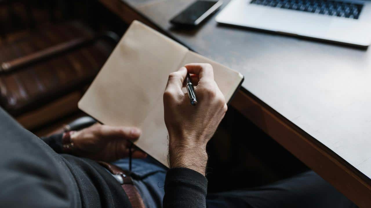 A person writing in a blank notebook at a desk with a laptop nearby.