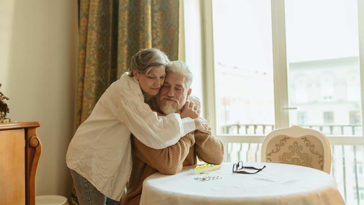 A senior woman hugging a seated senior man at a table near a window.
