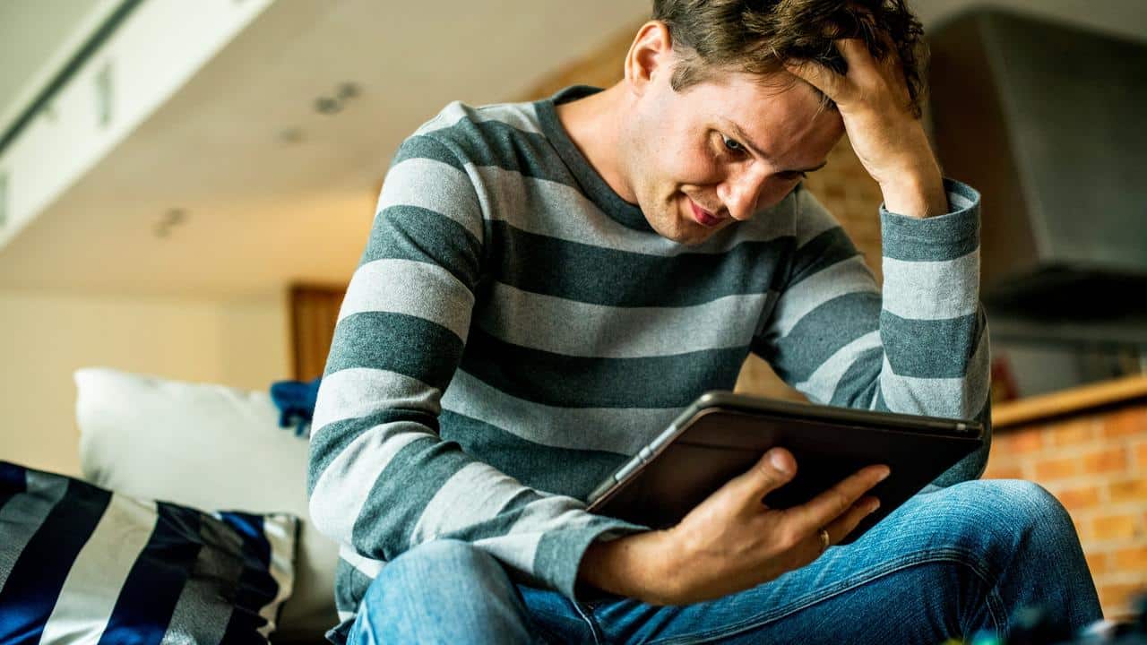A man sitting indoors and looking at a tablet with his hand on his head.