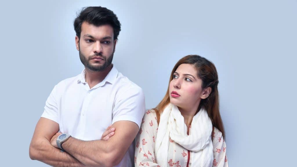 A man with crossed arms looks away while a woman looks at him with a frown.