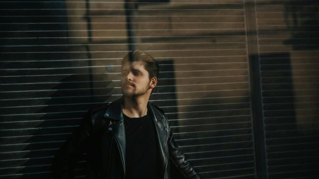 A man in a leather jacket standing by a window with blinds.