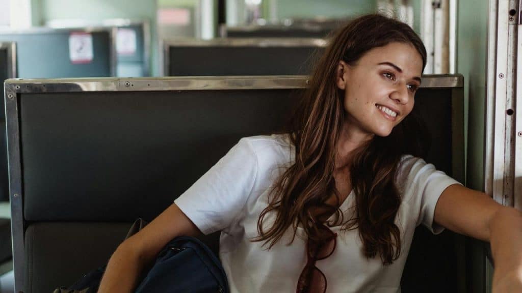 A smiling woman sitting by a train window with a bag beside her.