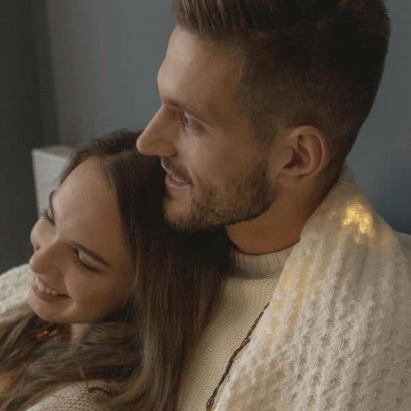 A couple cuddling under a blanket and smiling indoors.