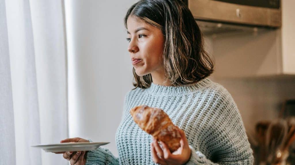 A woman holding a croissant and plate while looking out a window thoughtfully.