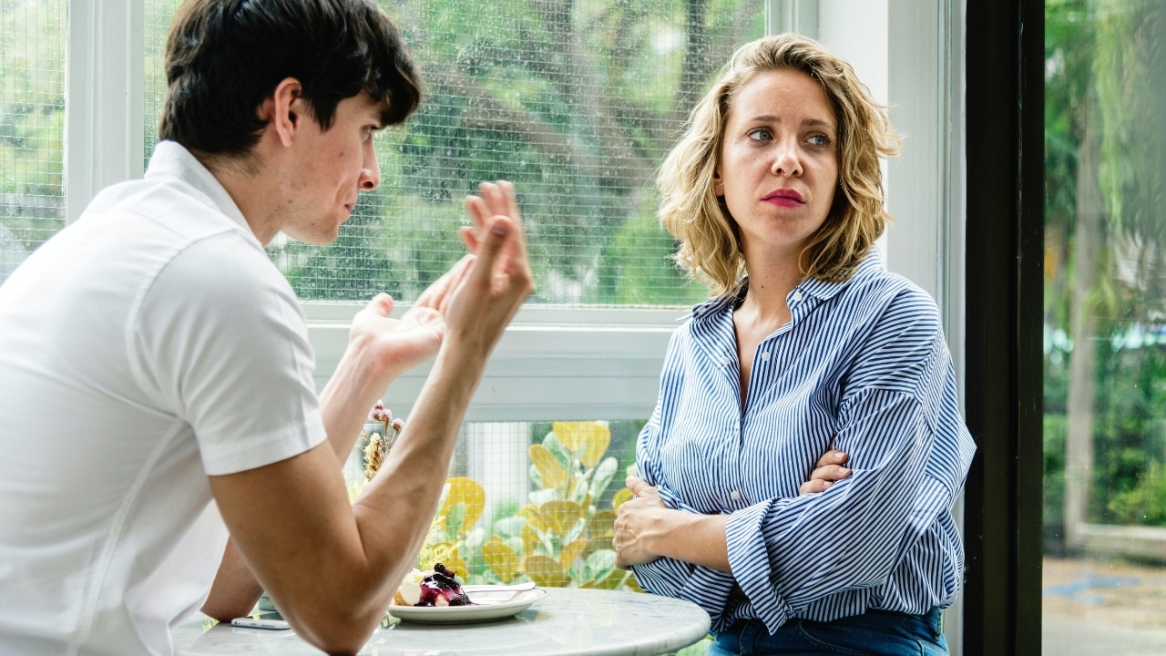 A woman with crossed arms looks away while a man gestures with his hands at a table.