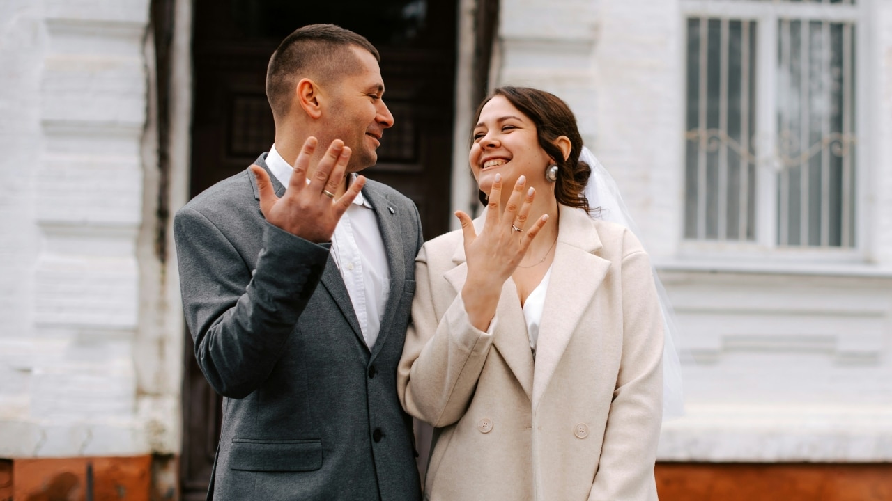 A smiling man and woman stand outside holding up their hands to show rings.