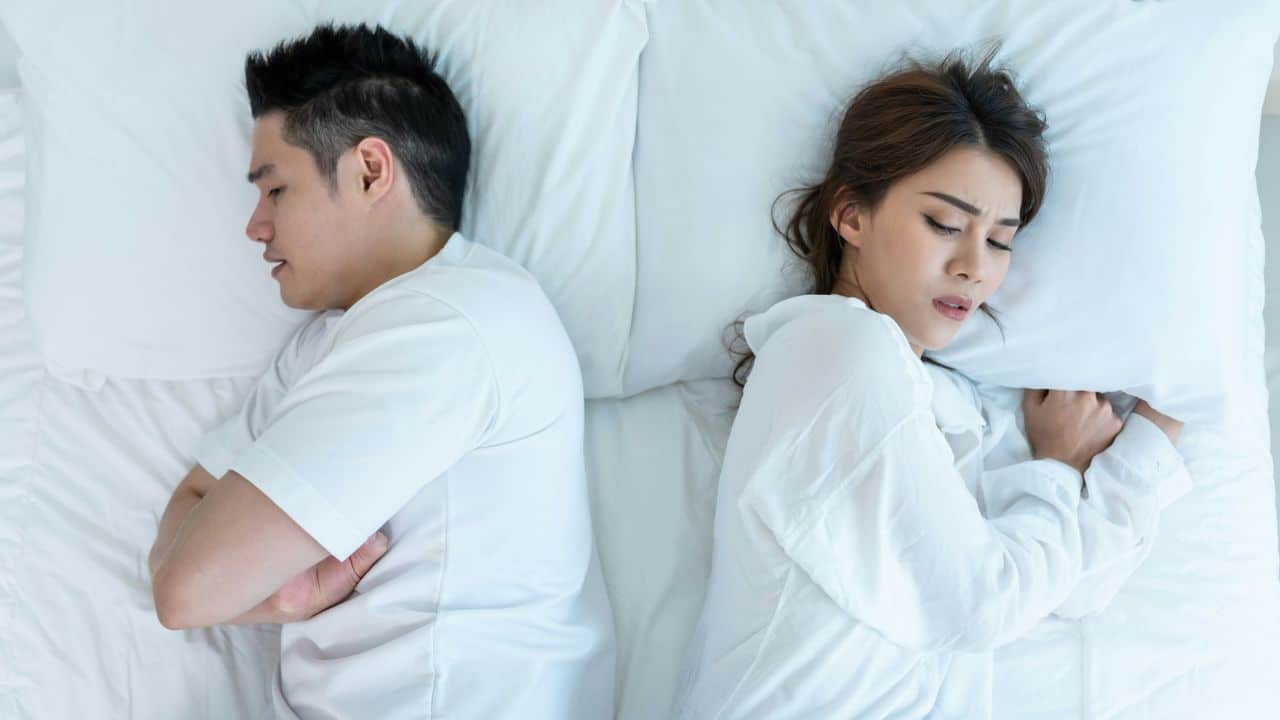 A man and woman lie in bed facing away from each other with eyes closed.